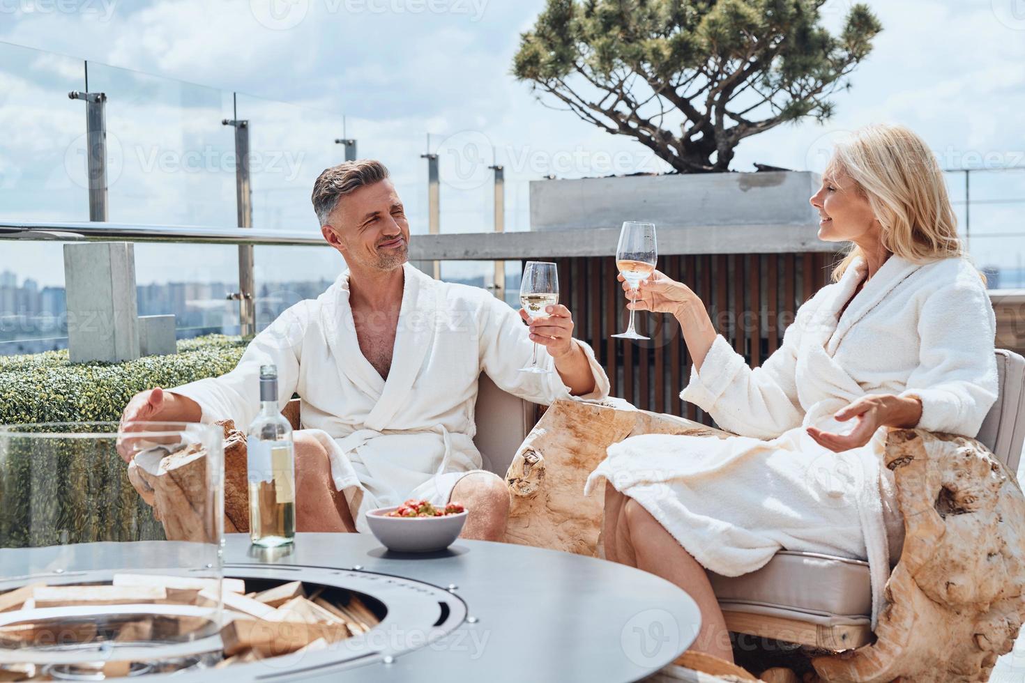 hermosa pareja madura en albornoces disfrutando de frutas y vino mientras se relaja en un hotel de lujo al aire libre foto