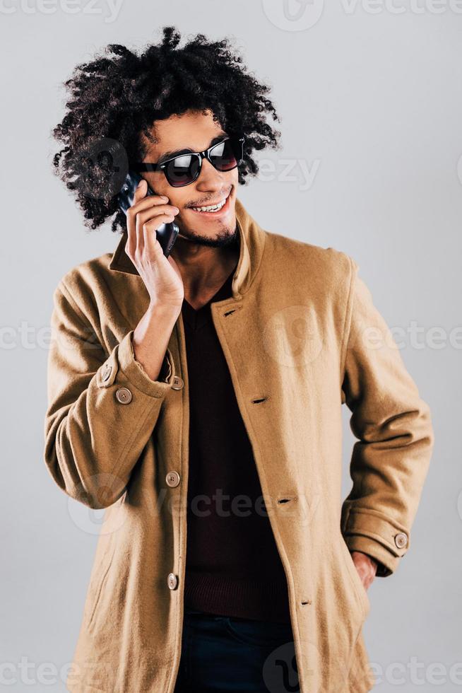 Phone call from friend. Cheerful young African man in sunglasses talking on mobile phone with smile while standing against grey background photo