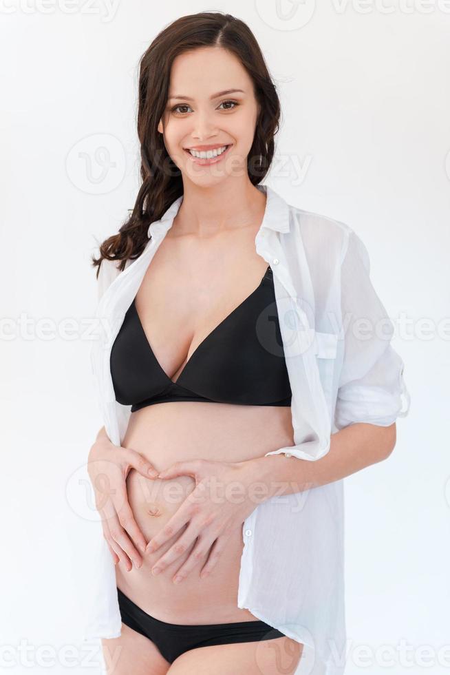 My baby will get a lot of love. Happy pregnant woman holding hands in heart shape on her abdomen and smiling while standing against white background photo