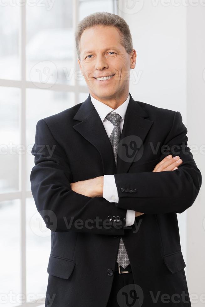 hombre de negocios confiado. hombre maduro alegre en ropa formal sonriendo a la cámara mientras está de pie cerca de la ventana foto