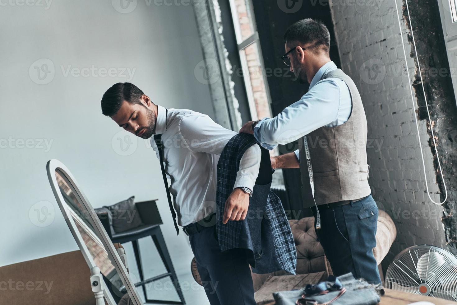 Suit fitting. Young fashionable designer helping his client to get dressed while standing in his workshop photo