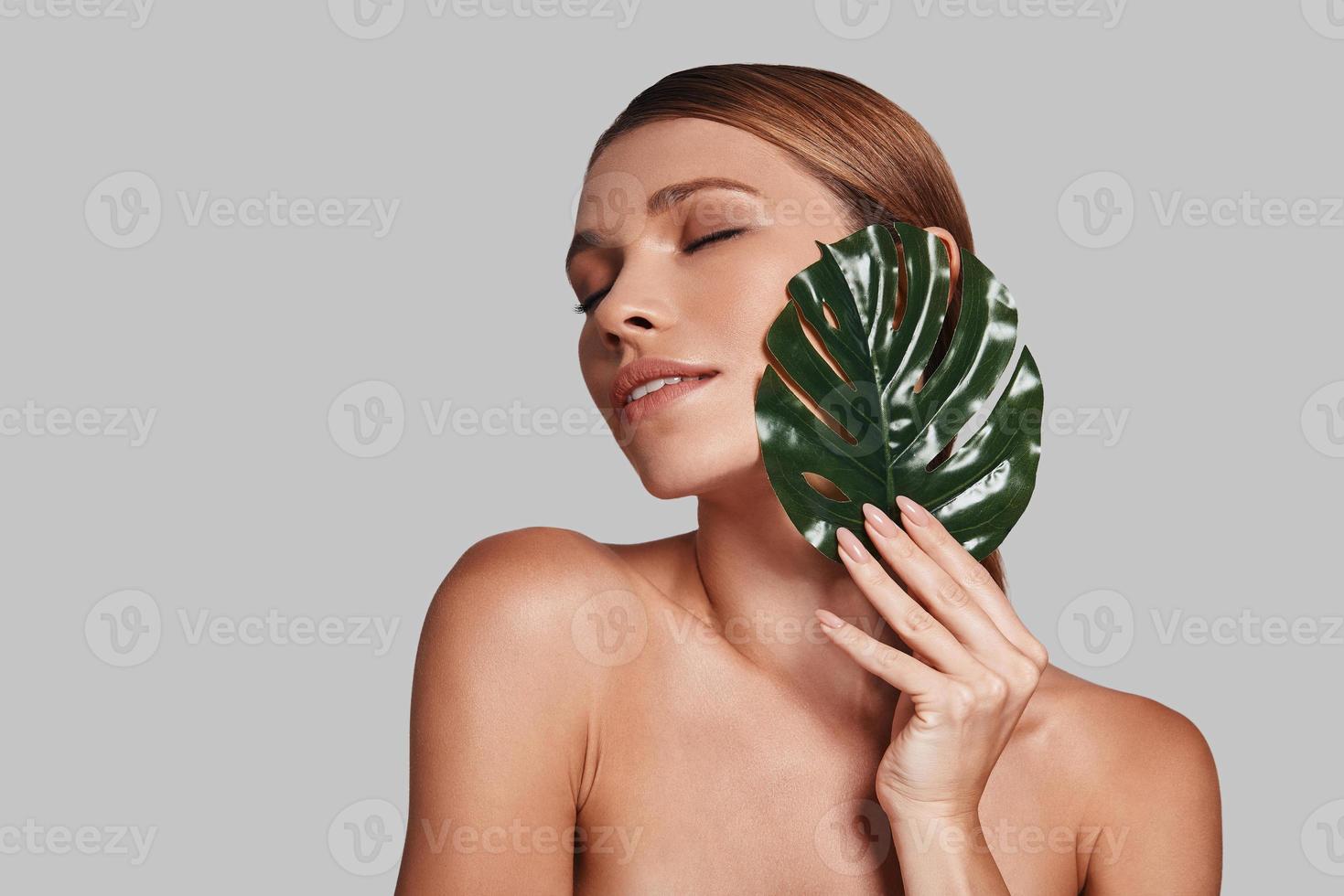Impossible to resist her beauty. Attractive young woman keeping eyes closed and covering with leaf while standing against grey background photo