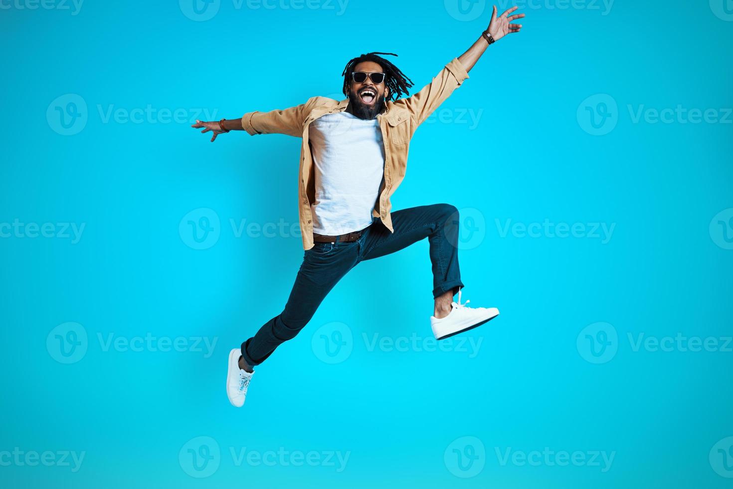 toda la longitud de un joven africano alegre con ropa informal gritando y gesticulando mientras se cierne sobre un fondo azul foto