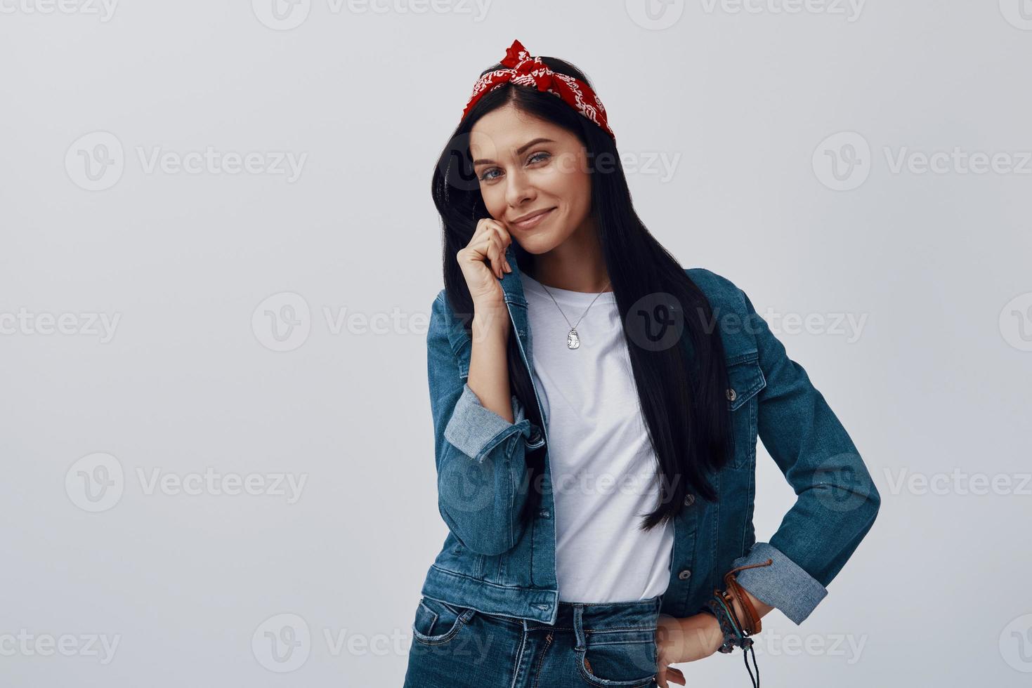 Attractive young woman in bandana looking at camera and smiling while standing against grey background photo
