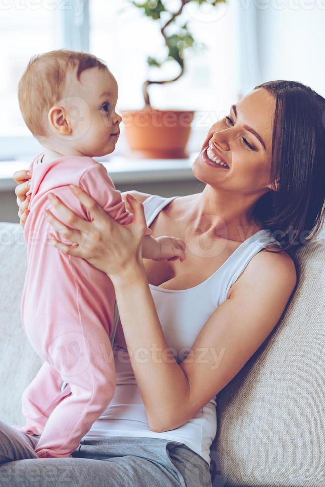 Simply admire her Cheerful beautiful young woman holding baby girl in her hands and looking at her with love while sitting on the couch at home photo