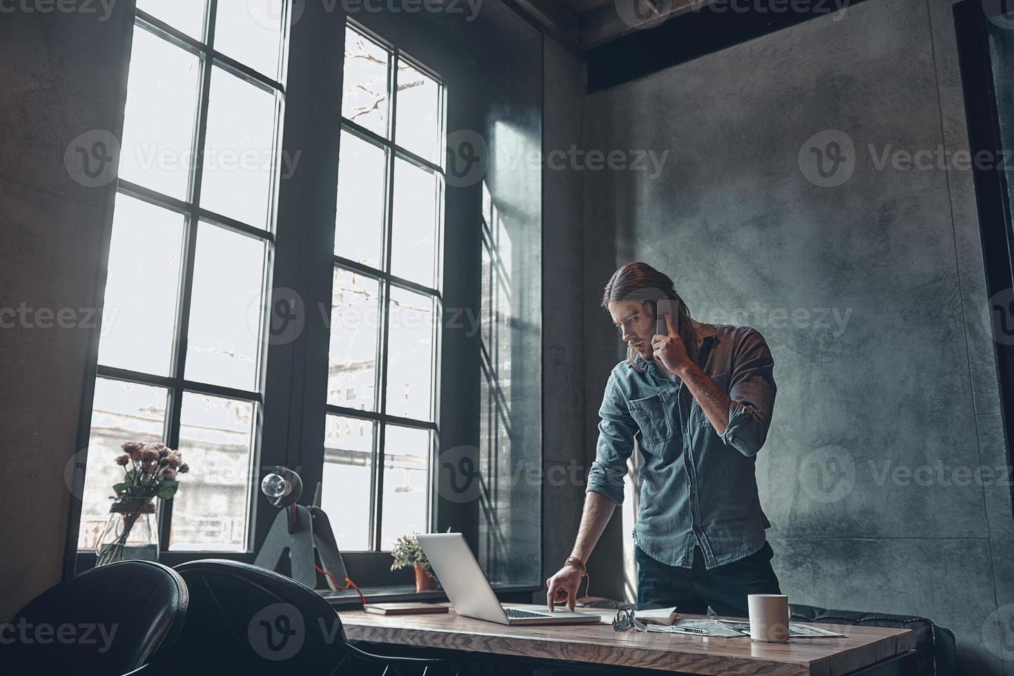 Developing new project using technologies. Serious young man talking on the mobile phone and working using laptop while standing near the office desk photo