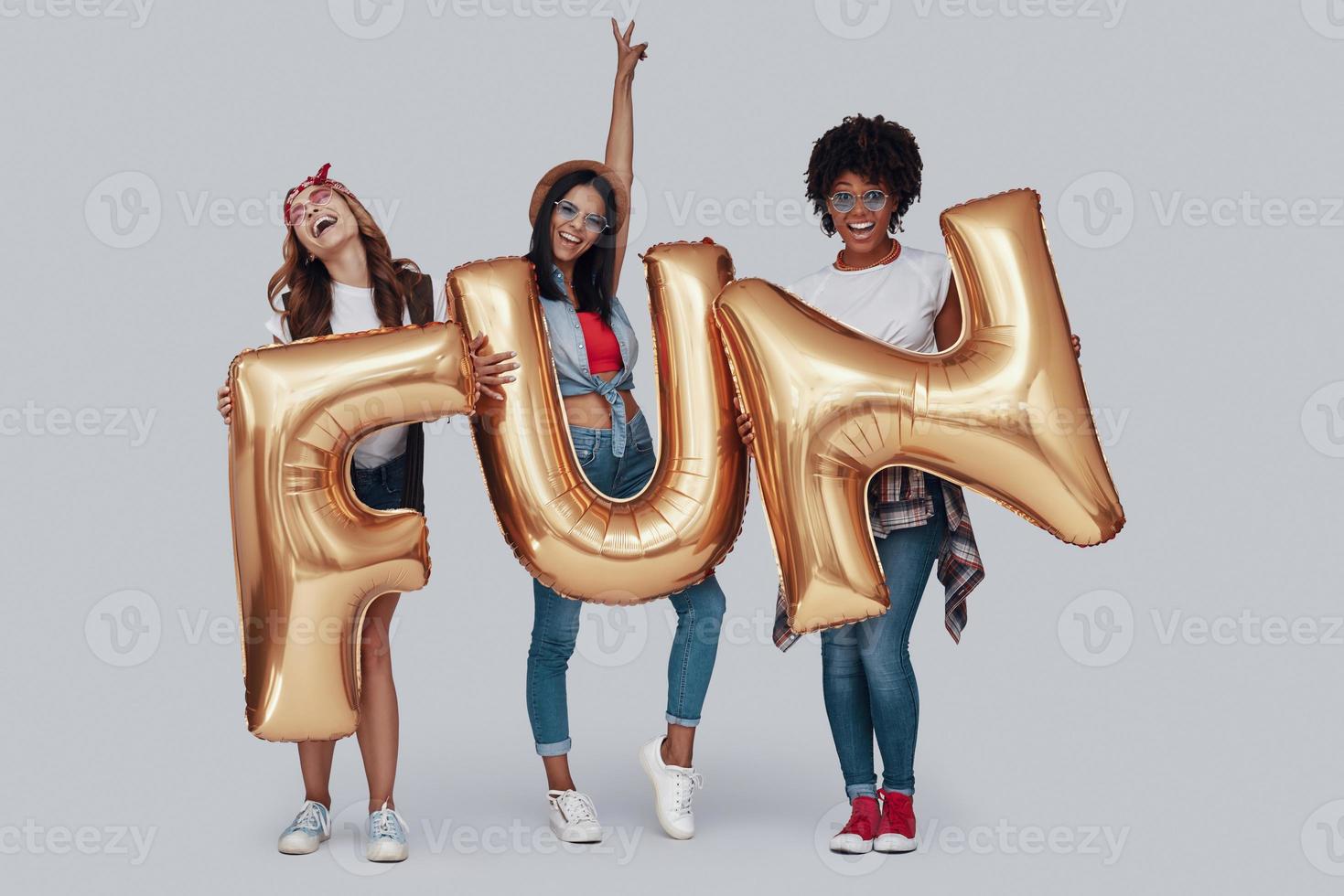 Full length of three attractive young women carrying balloons and smiling while standing against grey background photo