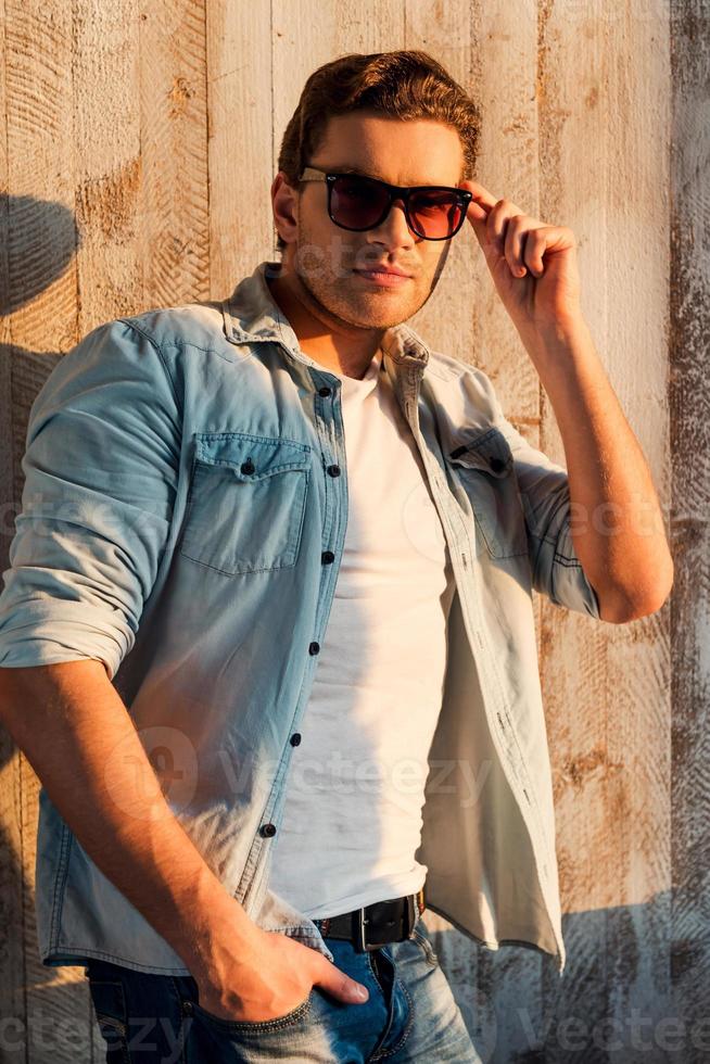 Cool and handsome. Handsome young man adjusting his sunglasses and leaning at the wall photo