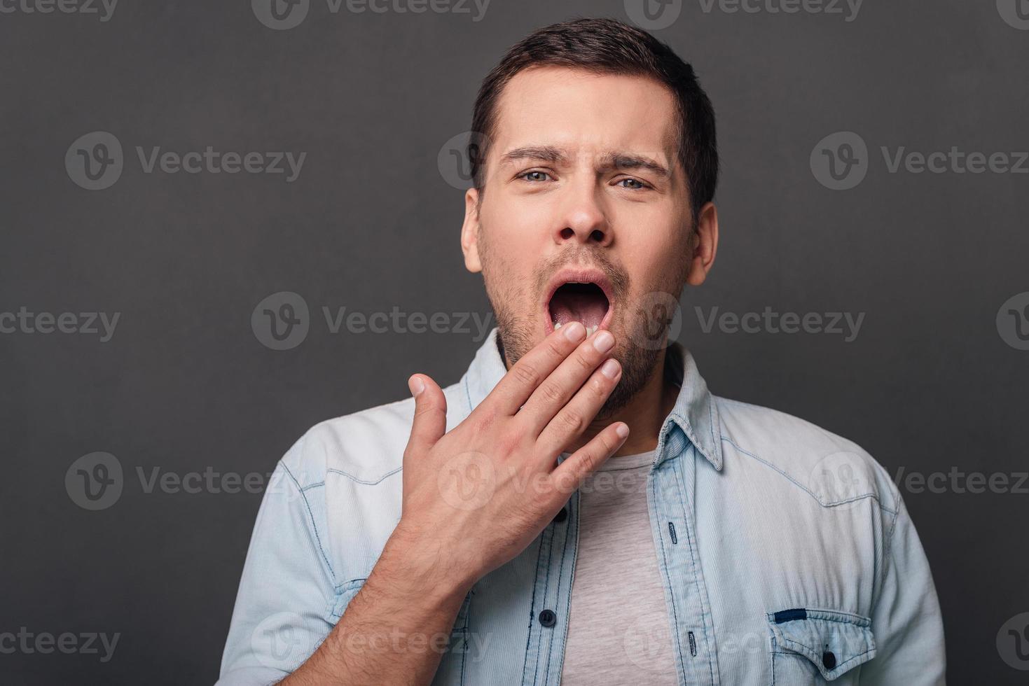 So bored Young handsome man covering mouth by hand and yawning while standing against grey background photo