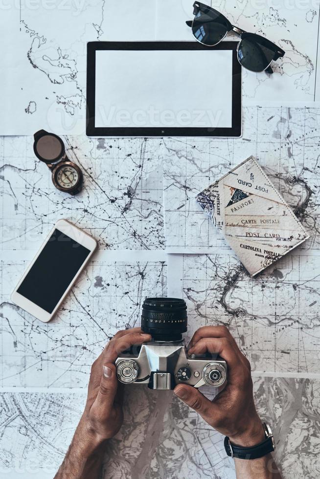 Real traveler. Close up top view of man holding photo camera with smart phone, sunglasses, compass and passport lying on map around