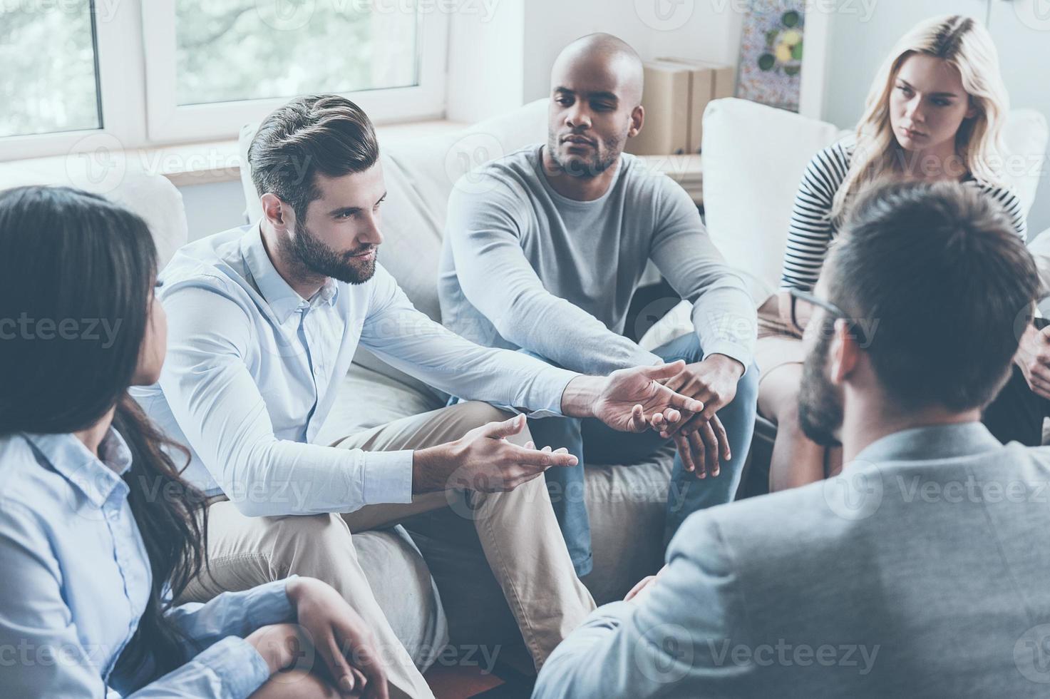 Sharing his problems with group. Group of young people sitting in circle while one man telling something and gesturing photo