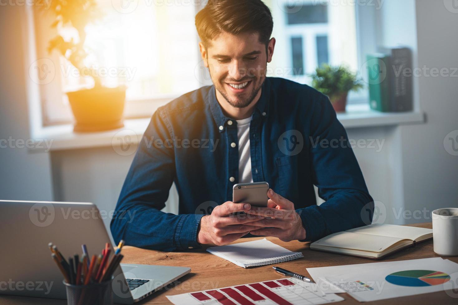 You have new message Cheerful young handsome man using his smartphone with smile while sitting at his working place photo