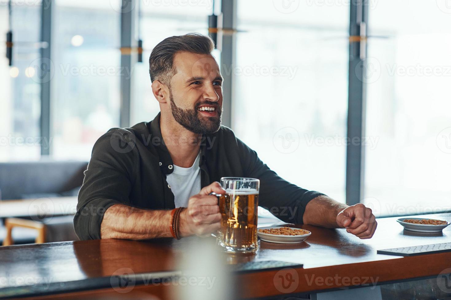 Cheerful young man in casual clothing enjoying beer while spending time in the pub photo