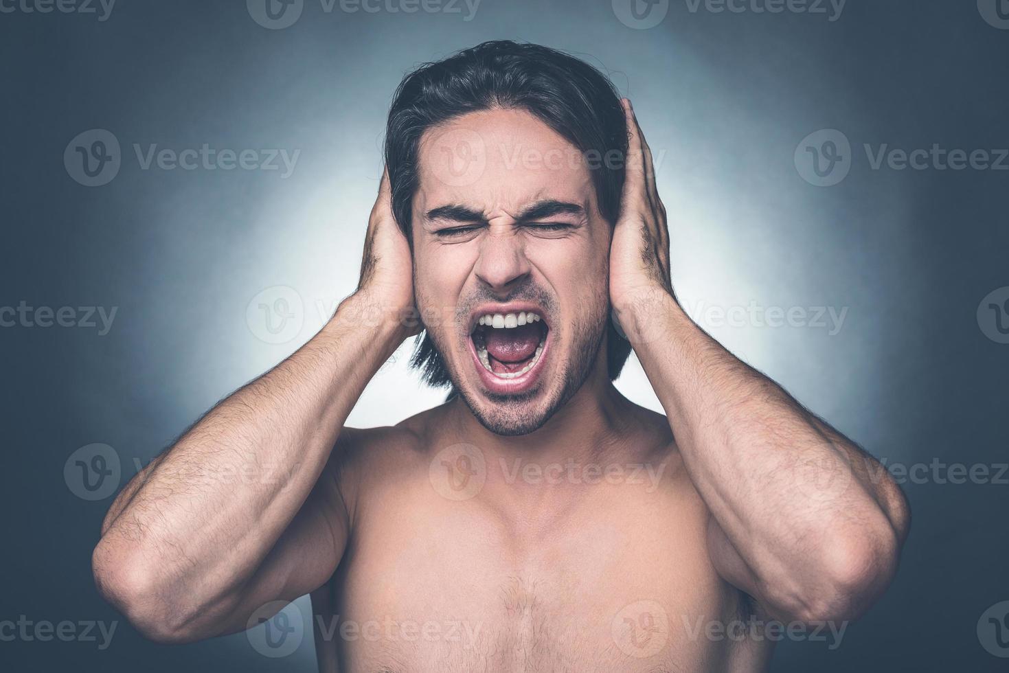 Too loud sound. Portrait of frustrated young shirtless man keeping eyes closed and covering ears with hands while standing against grey background photo