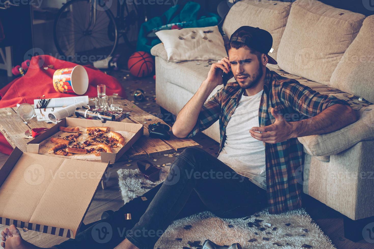 Handsome young man talking on mobile phone and gesturing while sitting on the floor in messy room after party photo