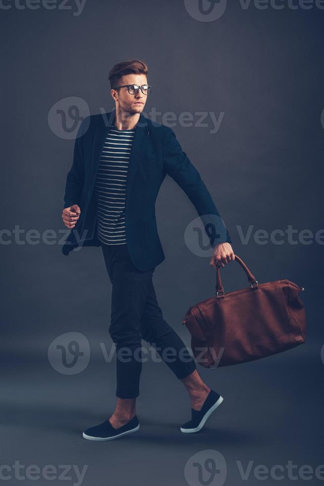 Style in action. Full length of confident young handsome man in glasses carrying bag and looking away while walking against grey background photo