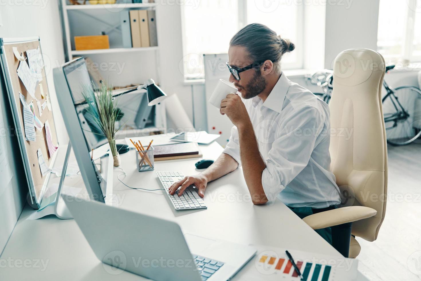 Putting ideas into something real. Thoughtful young man in shirt working using computer and drinking coffee while sitting in the office photo