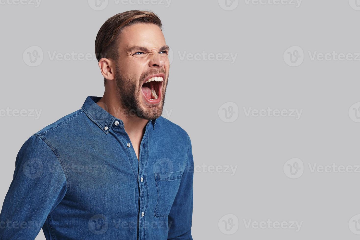 Furious man. Angry young man screaming while standing against grey background photo