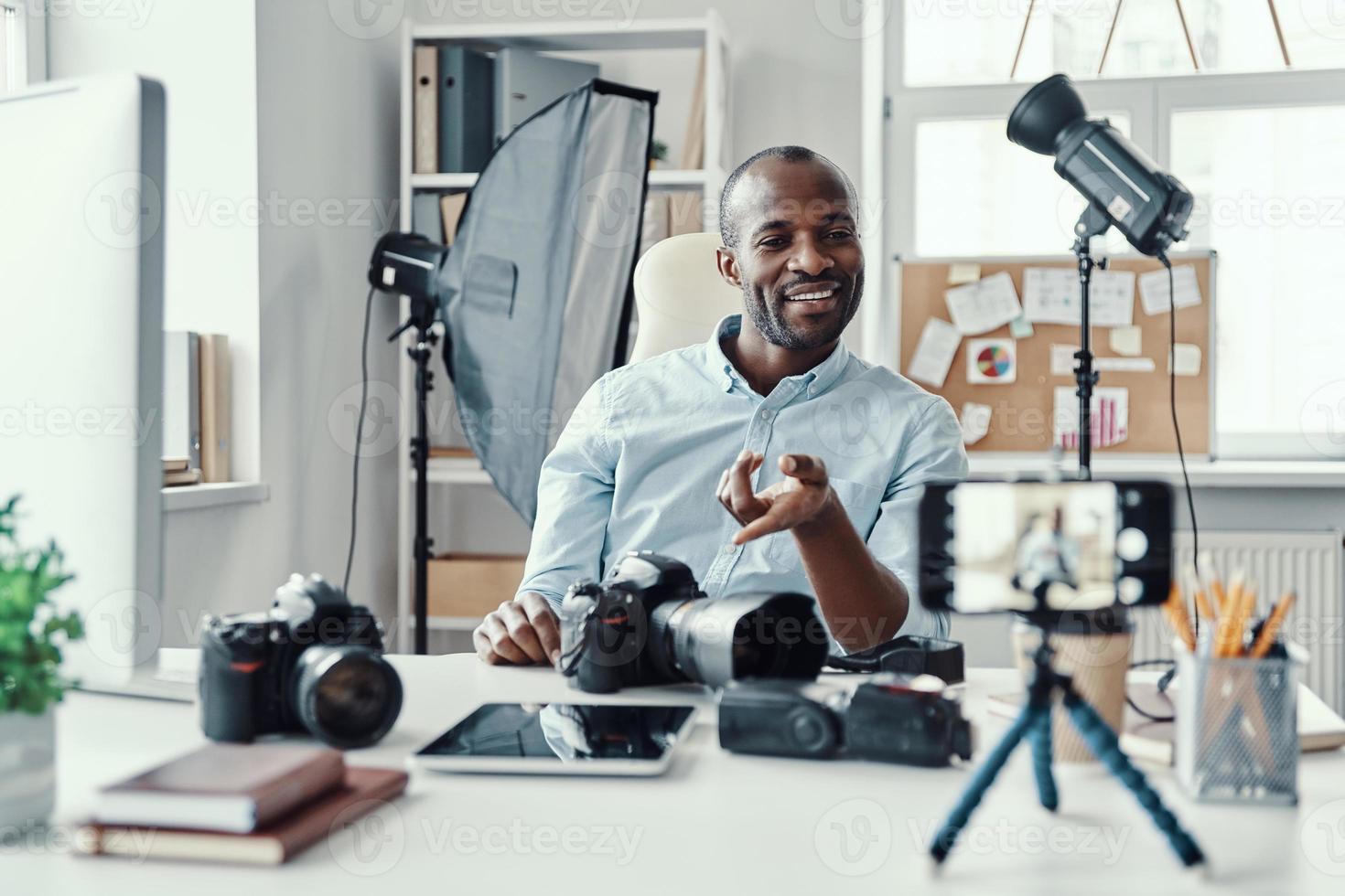 Happy young African man in shirt showing digital camera and telling something while making social media video photo
