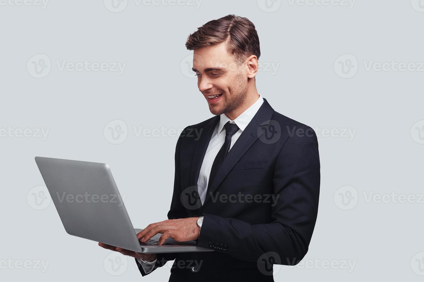 Ready to work hard. Good looking young man in full suit using laptop and smiling while standing against grey background photo