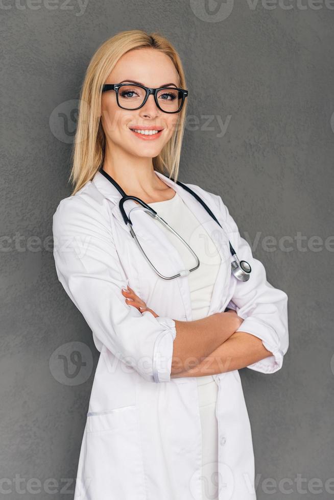 She can cure you. Beautiful young female doctor keeping arms crossed and looking at camera with smile while standing against grey background photo