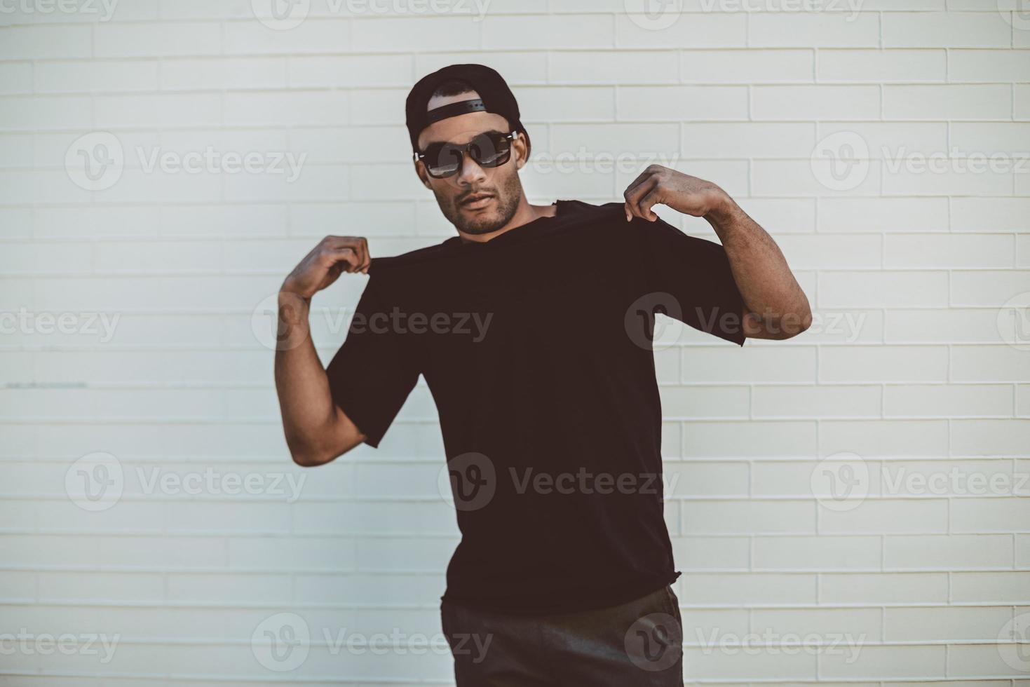 Feeling just casual. Handsome young African man touching his T-Shirt and looking at camera while standing in front of the brick wall outdoors photo