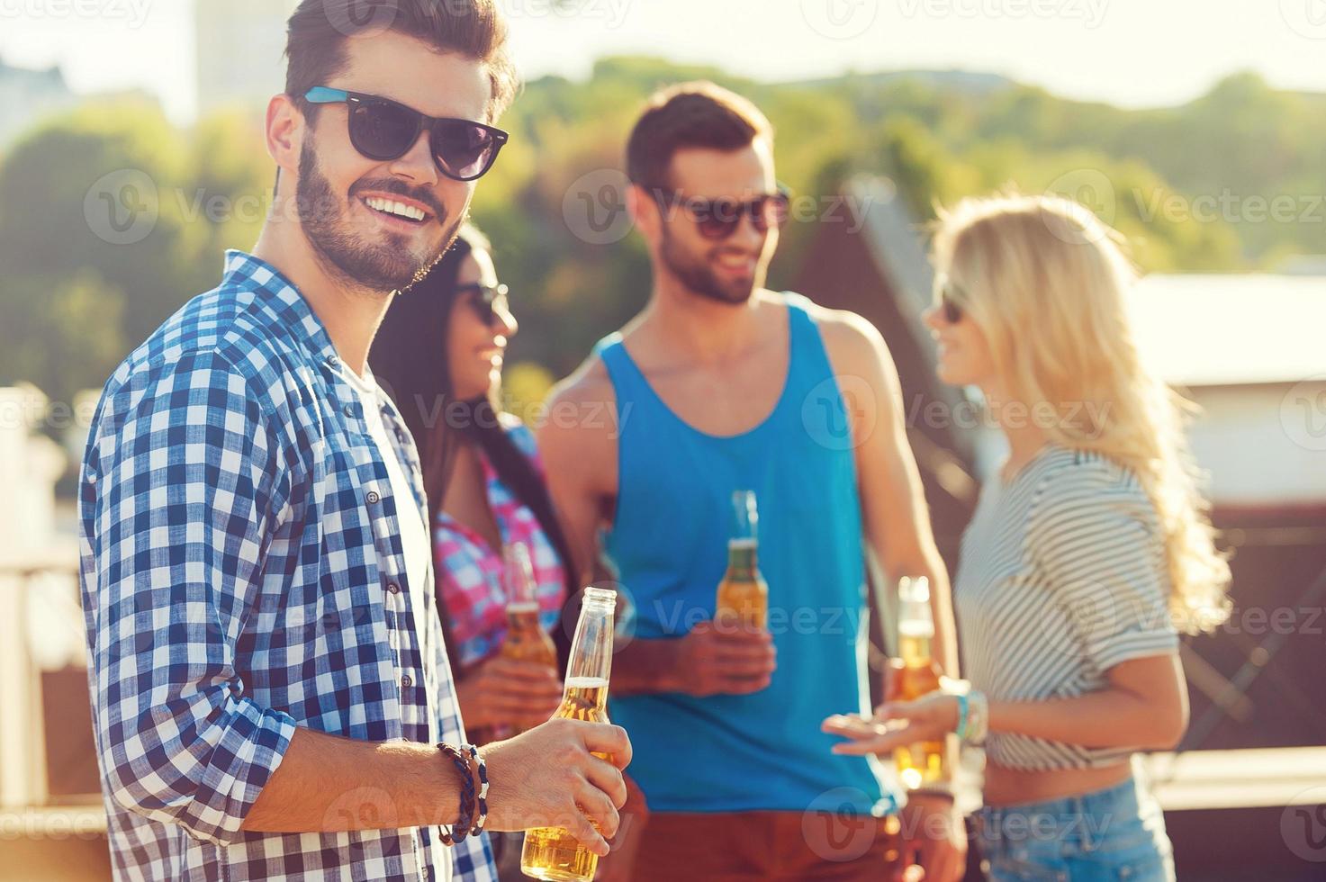 disfrutando de la cerveza con amigos. joven feliz sosteniendo una botella de cerveza y mirando a la cámara mientras tres personas hablan entre sí en el fondo foto