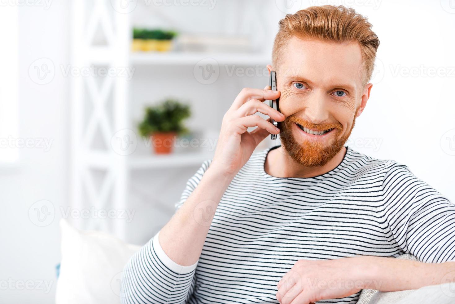 Lovely communication. Happy bearded man talking on the mobile phone and smiling at camera while sitting on sofa photo