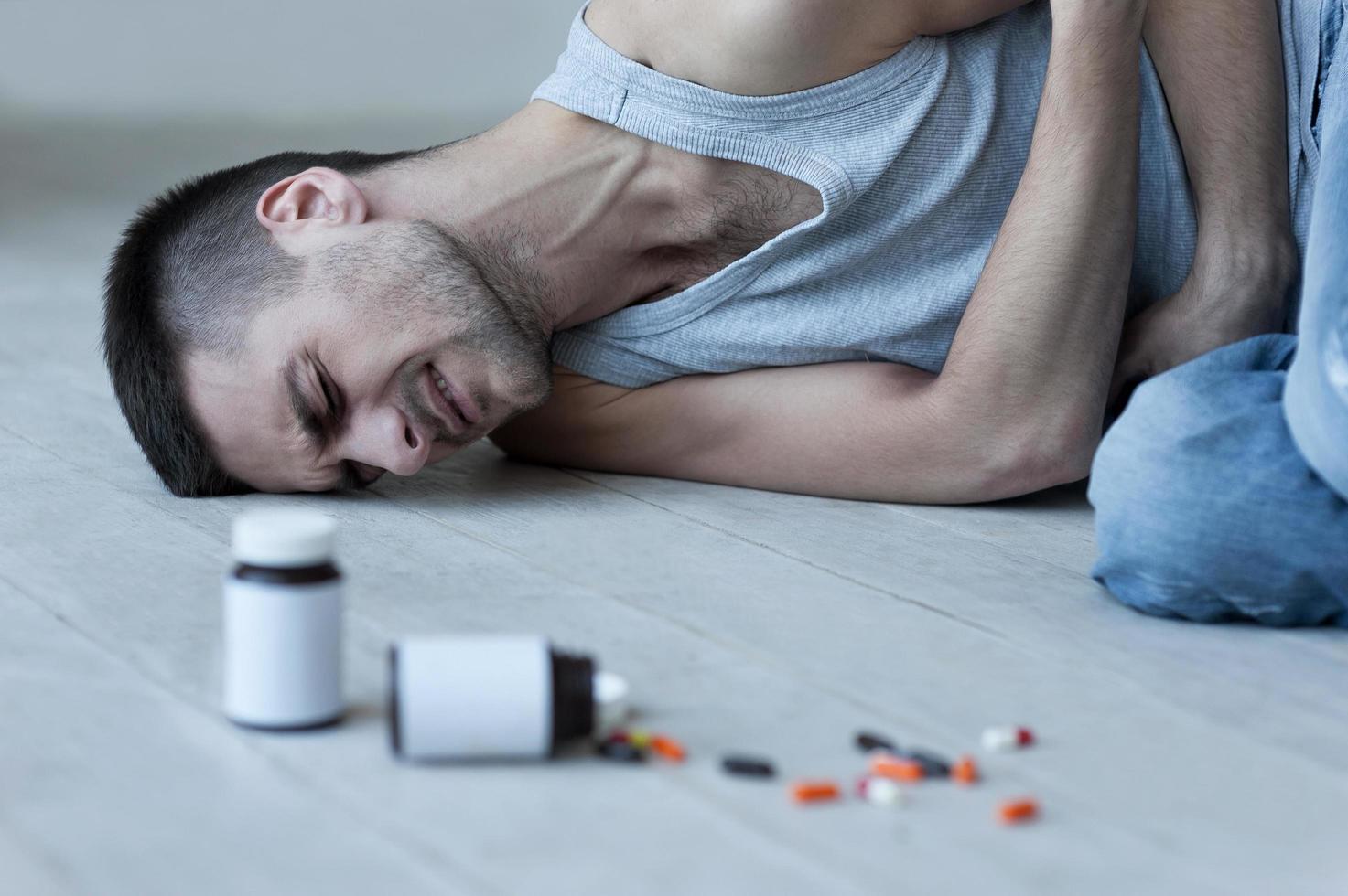 Numbing his pain. Young man lying on the floor and holding hands on stomach while bottles with pills laying near him photo