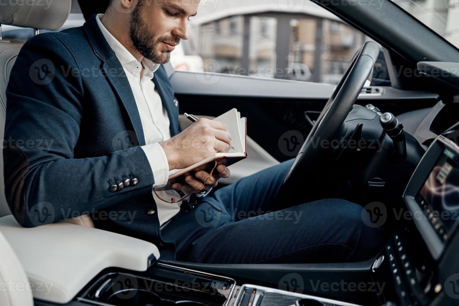 Planning the day. Handsome young man in full suit writing something down in personal organizer while sitting inside of the car photo