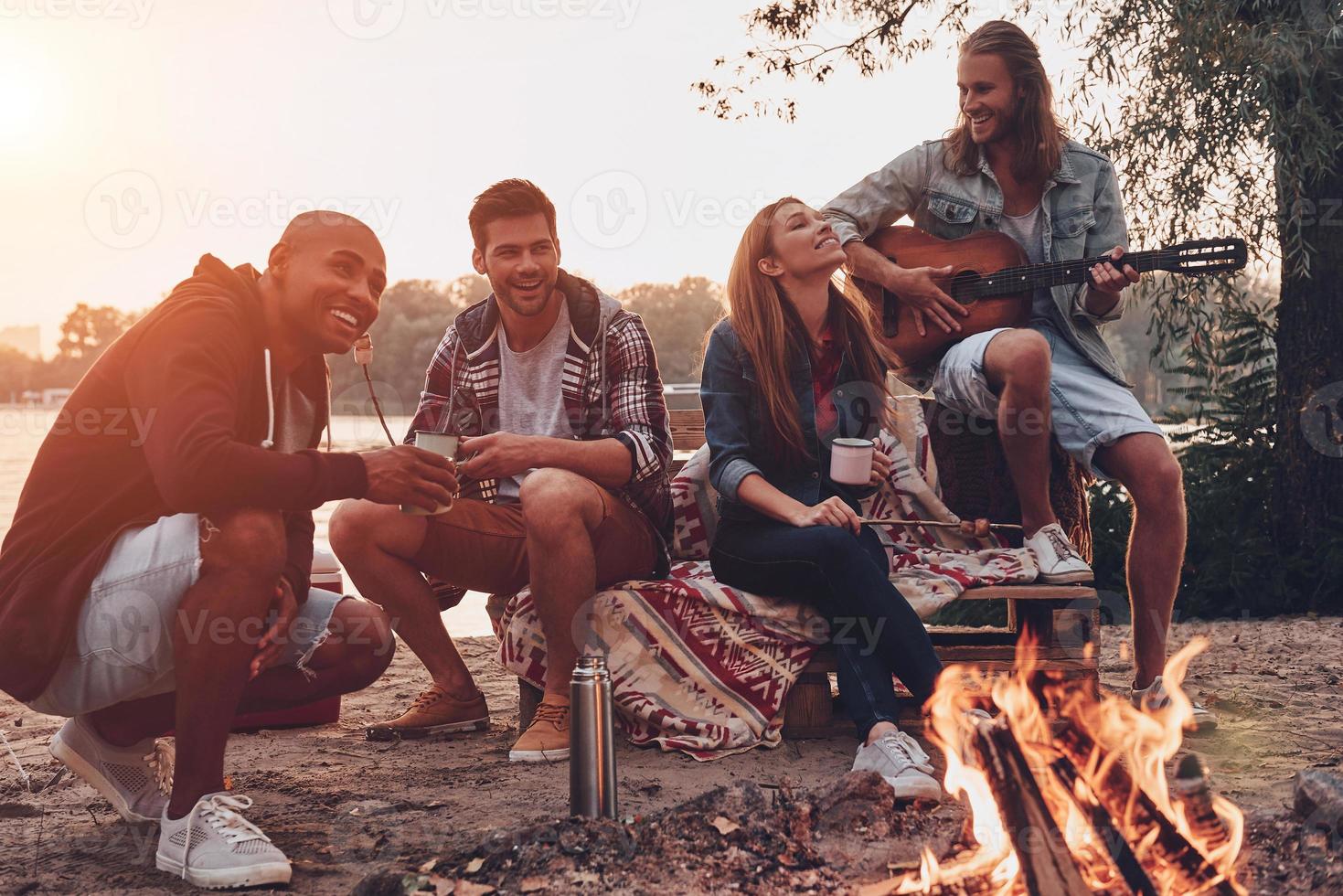 Carefree camping. Group of young people in casual wear smiling while enjoying beach party near the campfire photo