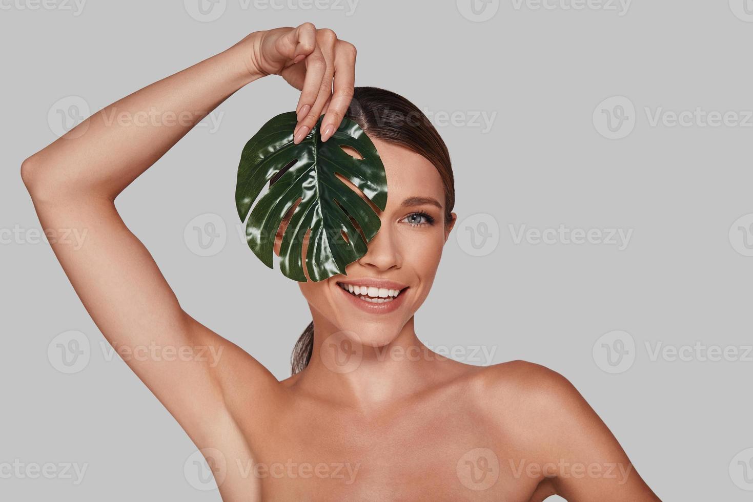 Happiness highlights her beauty. Attractive young woman looking at camera and covering face with leaf while standing against grey background photo