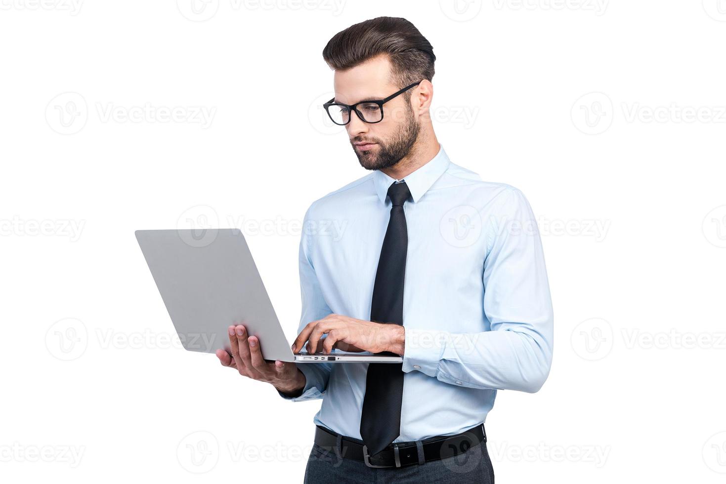 Concentrated on work. Confident young handsome man in shirt and tie working on laptop while standing against white background photo