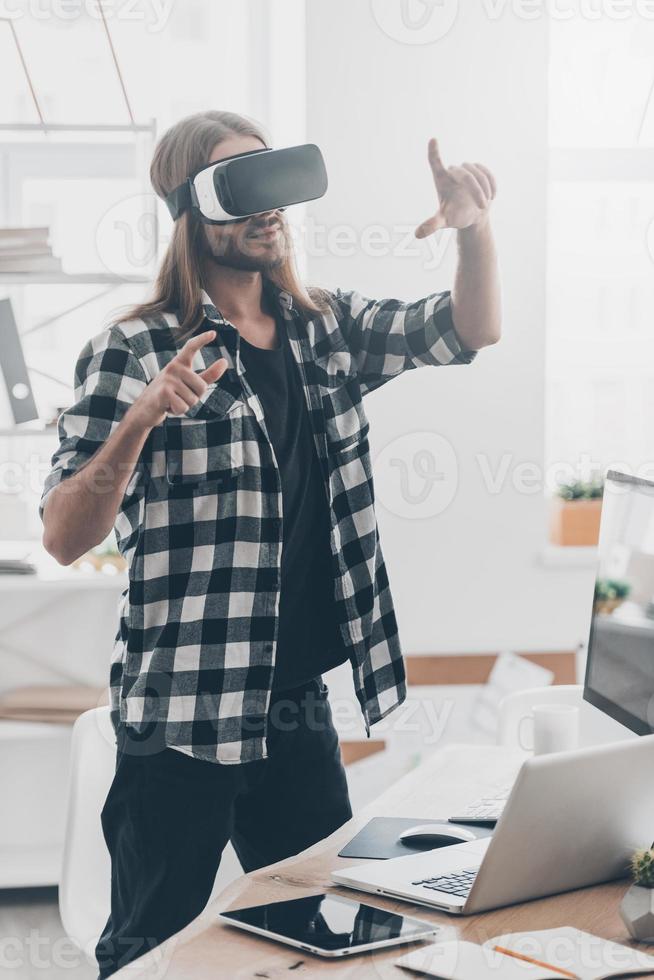 Enjoying new reality. Handsome young man with long hair in virtual reality headset gesturing while standing in creative office photo