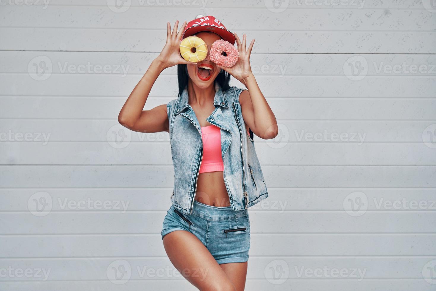 ella no sabe lo que la aburre. mujeres jóvenes juguetonas sosteniendo donuts contra sus ojos y sonriendo mientras están de pie contra la puerta del garaje foto