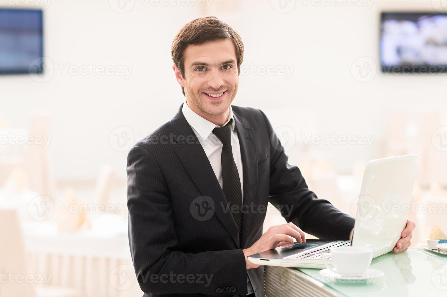 I am always connected. Handsome young man in formalwear using his laptop and smiling at camera while standing at the bar photo