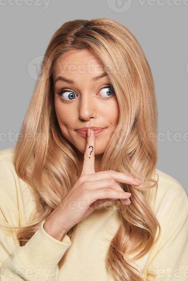 What if Beautiful young woman keeping index finger on her lips while standing against grey background photo