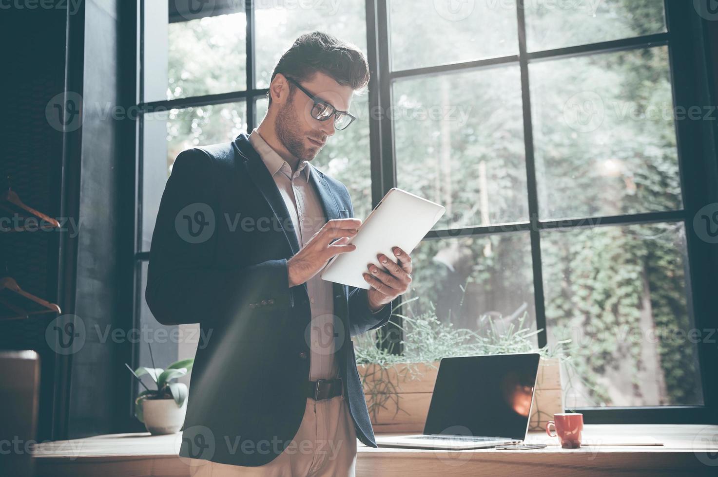 Working on new tablet. Low angle view of confident young man working on digital tablet while standing in front of the big window in office or cafe photo