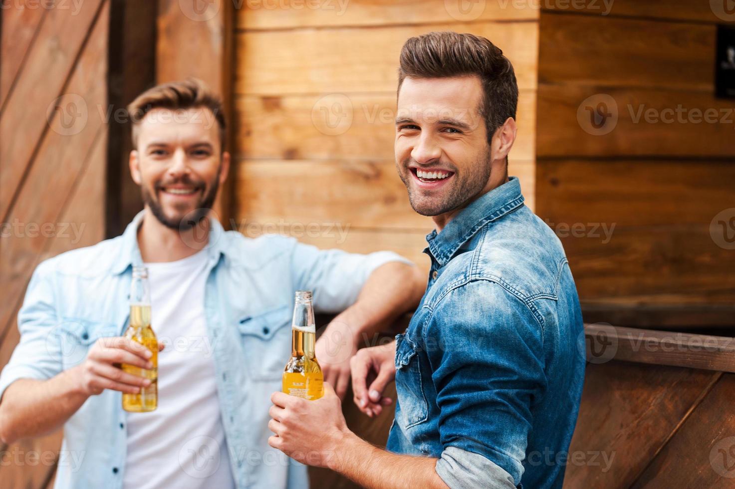 reunión de viejos amigos. dos jóvenes alegres sosteniendo botellas con cerveza y mirando a la cámara mientras se inclinan en el mostrador del bar foto