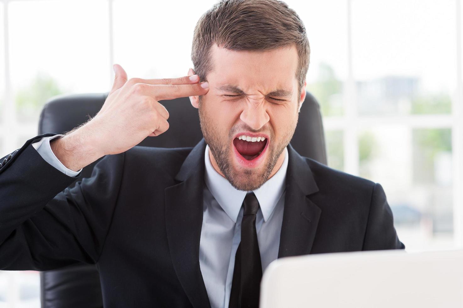 Emotional stress. Furious young man in formalwear touching his temple with finger gun and keeping eyes closed while sitting at his working place photo