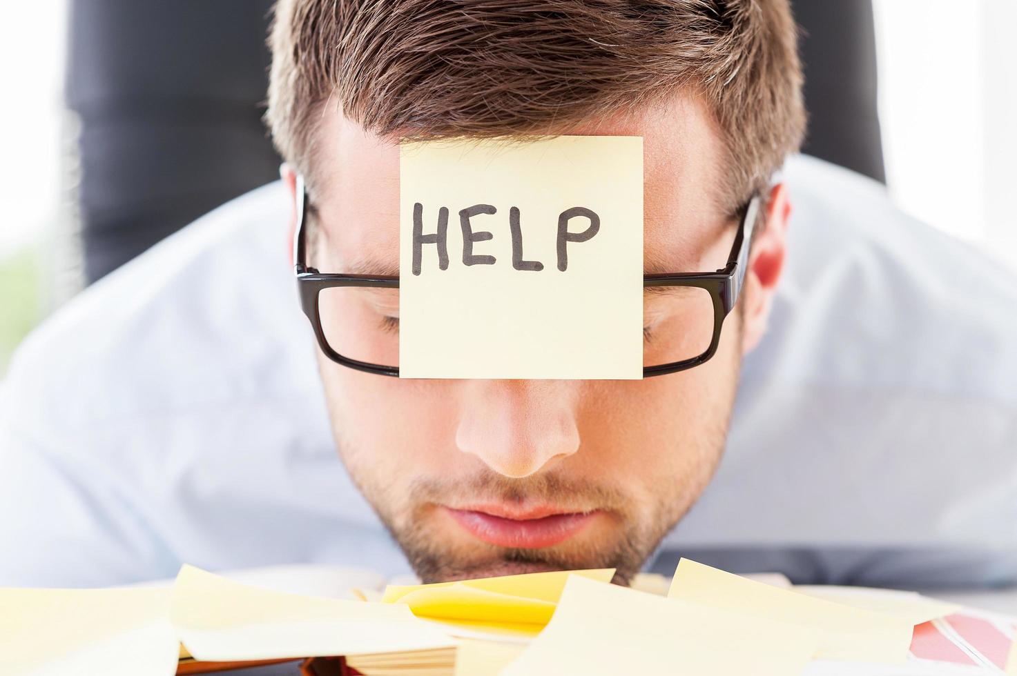 He needs help. Frustrated young man in formalwear with adhesive note on his forehead leaning his head at the table photo
