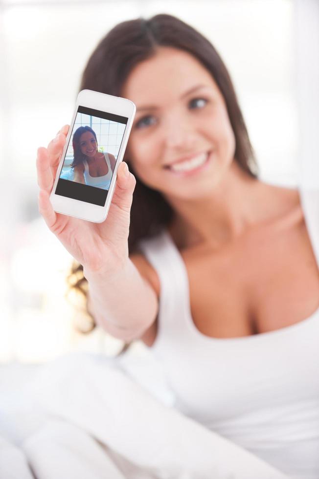 Making selfie. Beautiful young smiling woman sitting in bed and making self photos with mobile phone