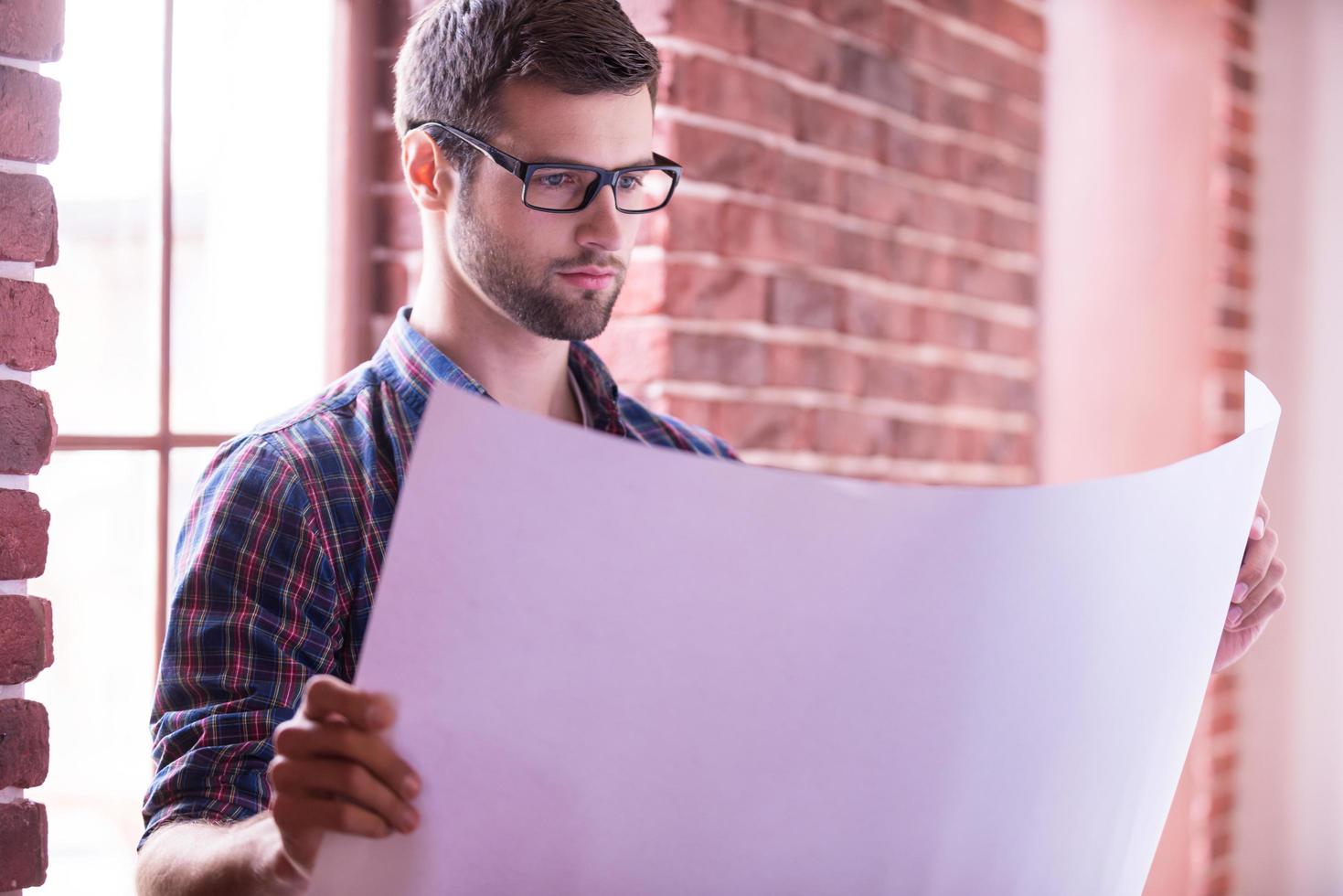 arquitecto examinando planos. joven confiado en anteojos examinando planos mientras está de pie cerca de la ventana foto