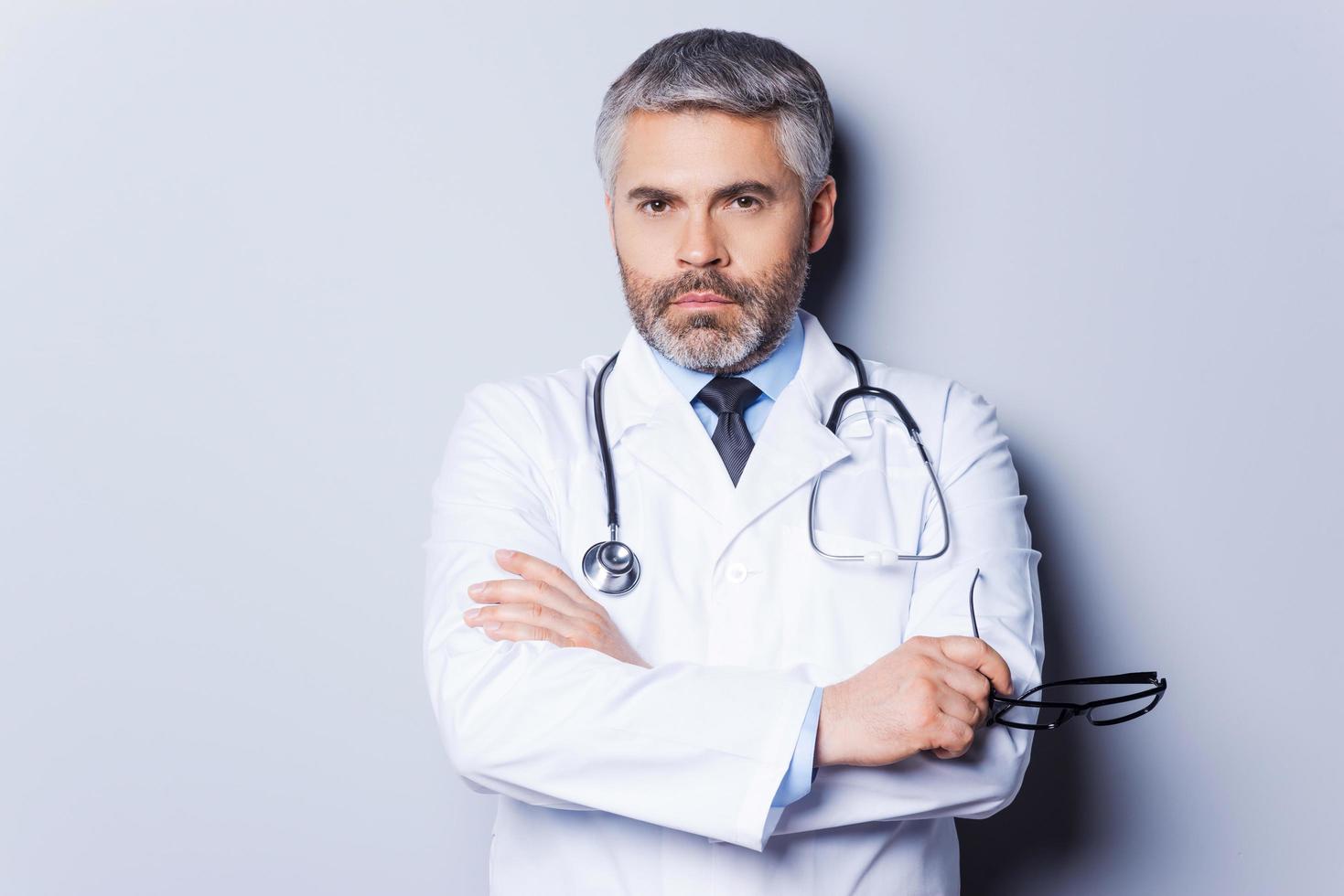 médico confiado. médico de pelo gris maduro mirando a la cámara y manteniendo los brazos cruzados mientras está de pie contra el fondo gris foto