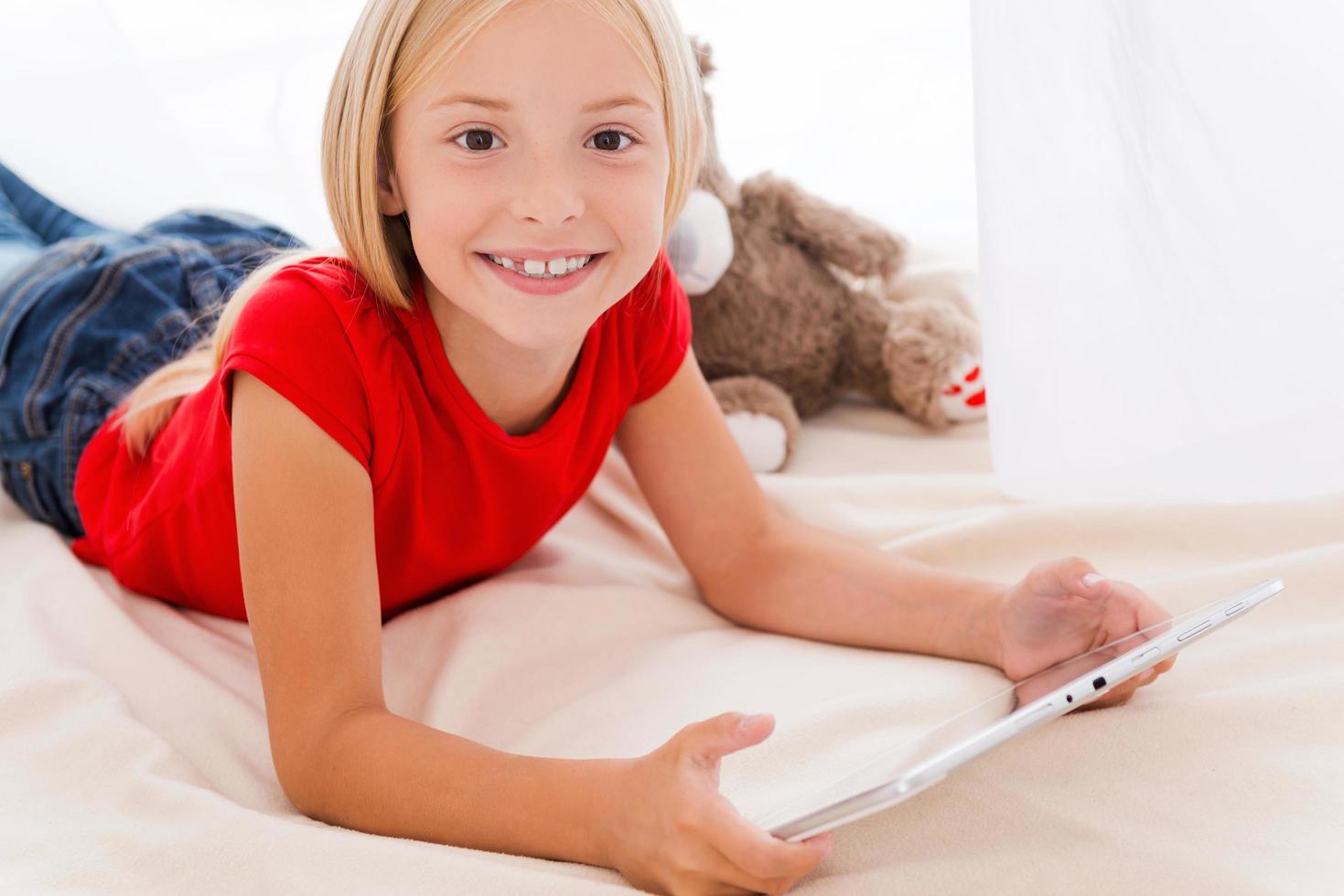 Technologies become easier. Cute little girl holding digital tablet and smiling while lying in bed photo