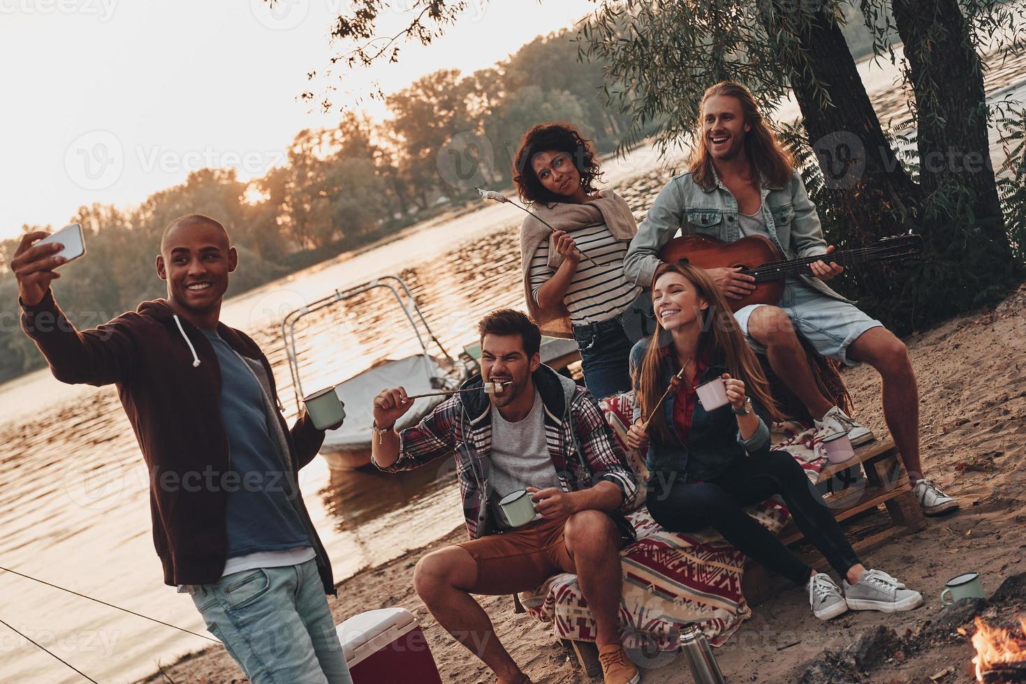 capturando momentos felices. grupo de jóvenes con ropa informal sonriendo y tomándose selfie mientras disfrutan de una fiesta en la playa cerca del lago foto
