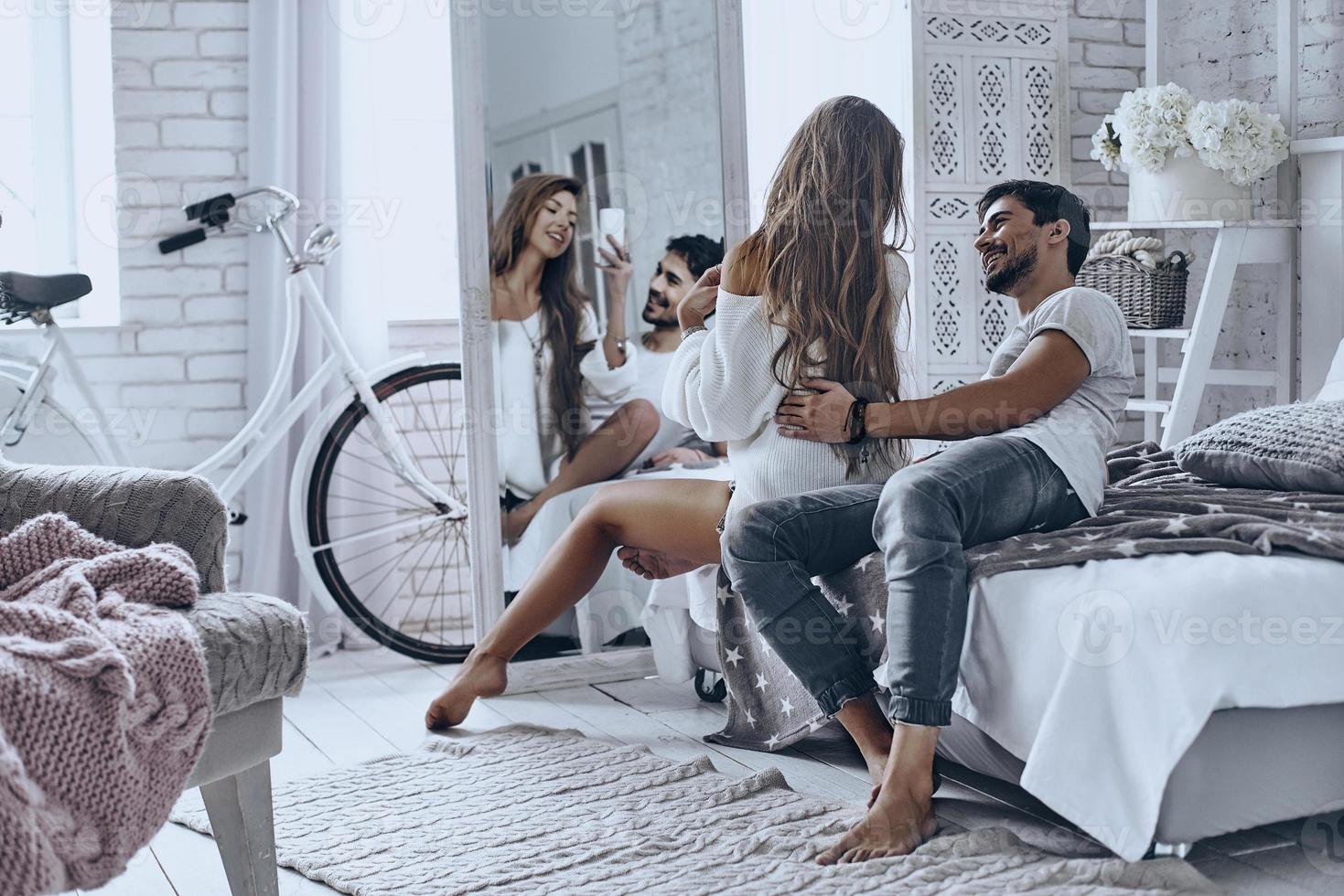 Reflection of love. Beautiful young couple resting at home and smiling while woman taking selfie in the mirror photo
