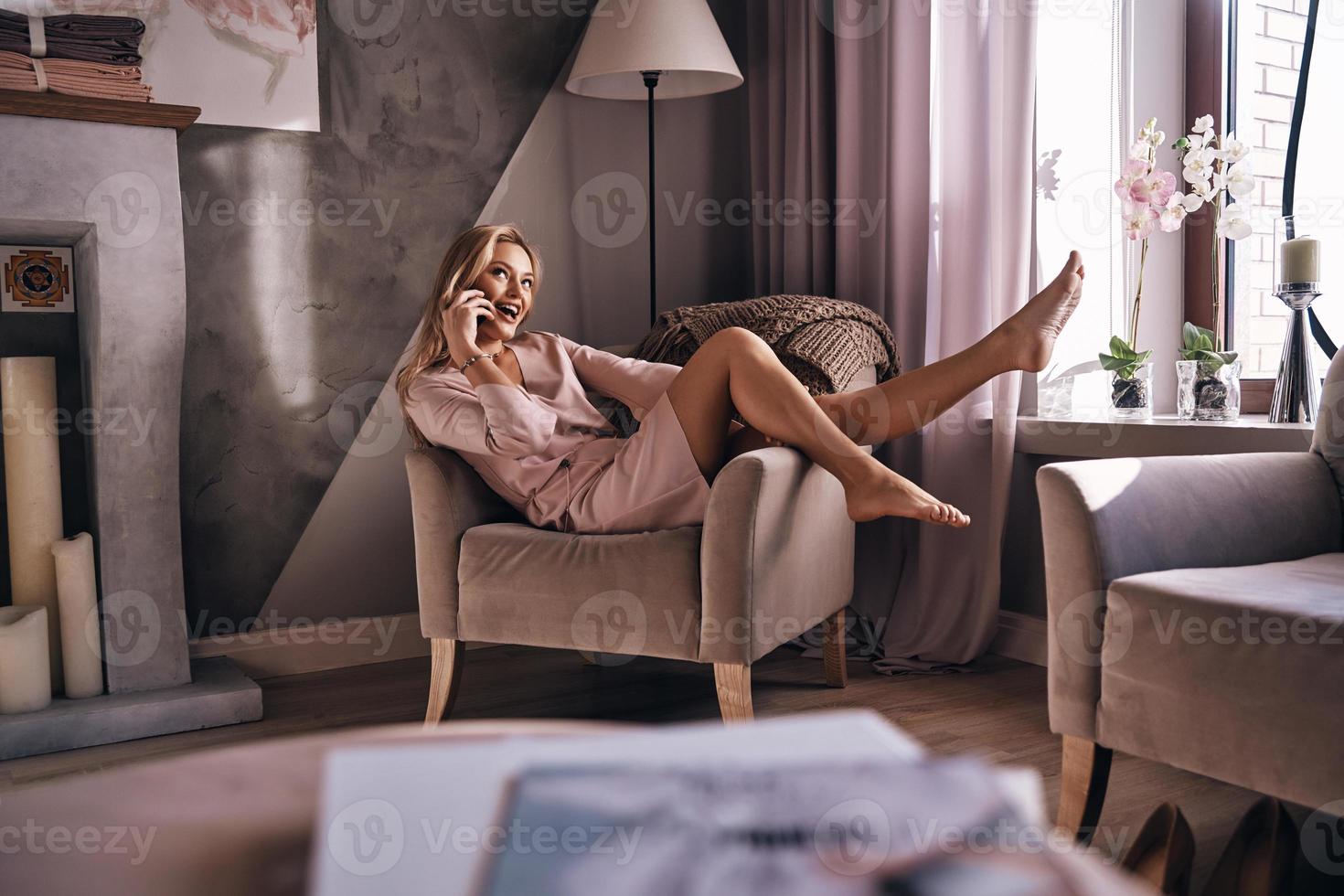 Happy to talk with friend.  Attractive young woman in elegant dress talking on the smart phone and smiling while sitting in the armchair photo