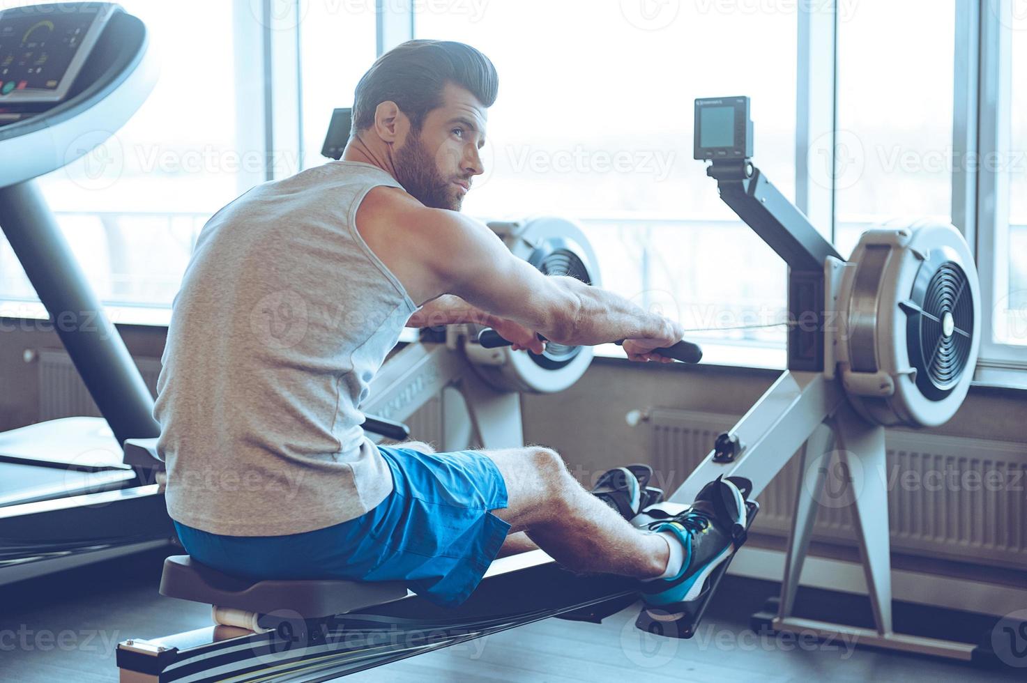 Rowing at gym. Side view of young man in sportswear doing rowing in front of window at gym and looking away photo