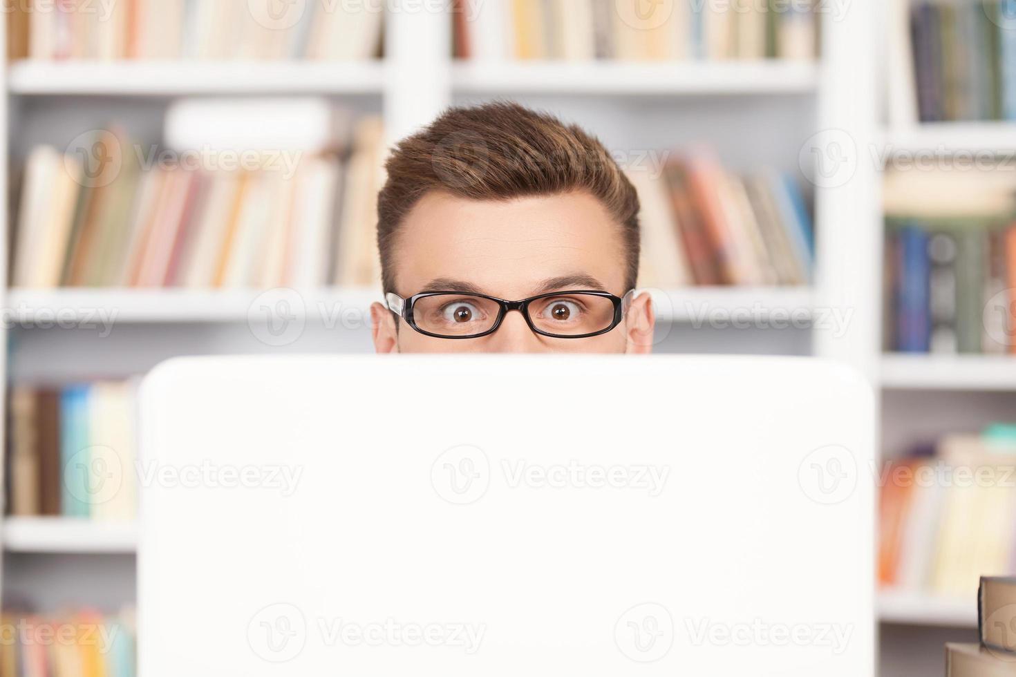 Shocking discovery. Shocked young nerd man in glasses looking out of computer while sitting at the library photo